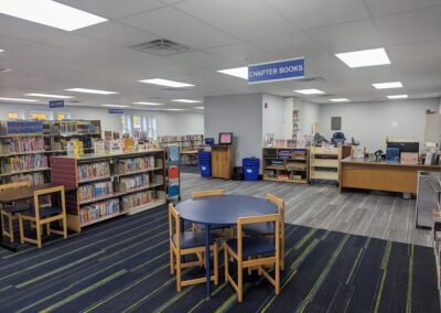 Shelving in the children's area of the library with tables and chairs. You can see the circulation desk in this photo as well.